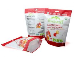 Three stand-up pouches for fruit candies. Two pouches are upright, and one is lying on its side, showing its hexagonal base. The pouches have a white base with red accents at the top and bottom. Graphics of fruit pieces and children are visible. Text includes 'YumEarth', 'freshest fruit', and 'organic hard candies'.