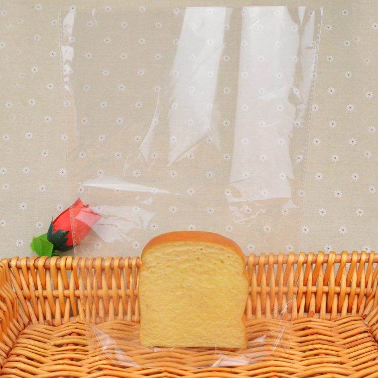 A slice of bread is placed inside a clear, flat food packaging bag. The bag is open at the top and rests on a woven basket. A red rose decoration is visible to the left.