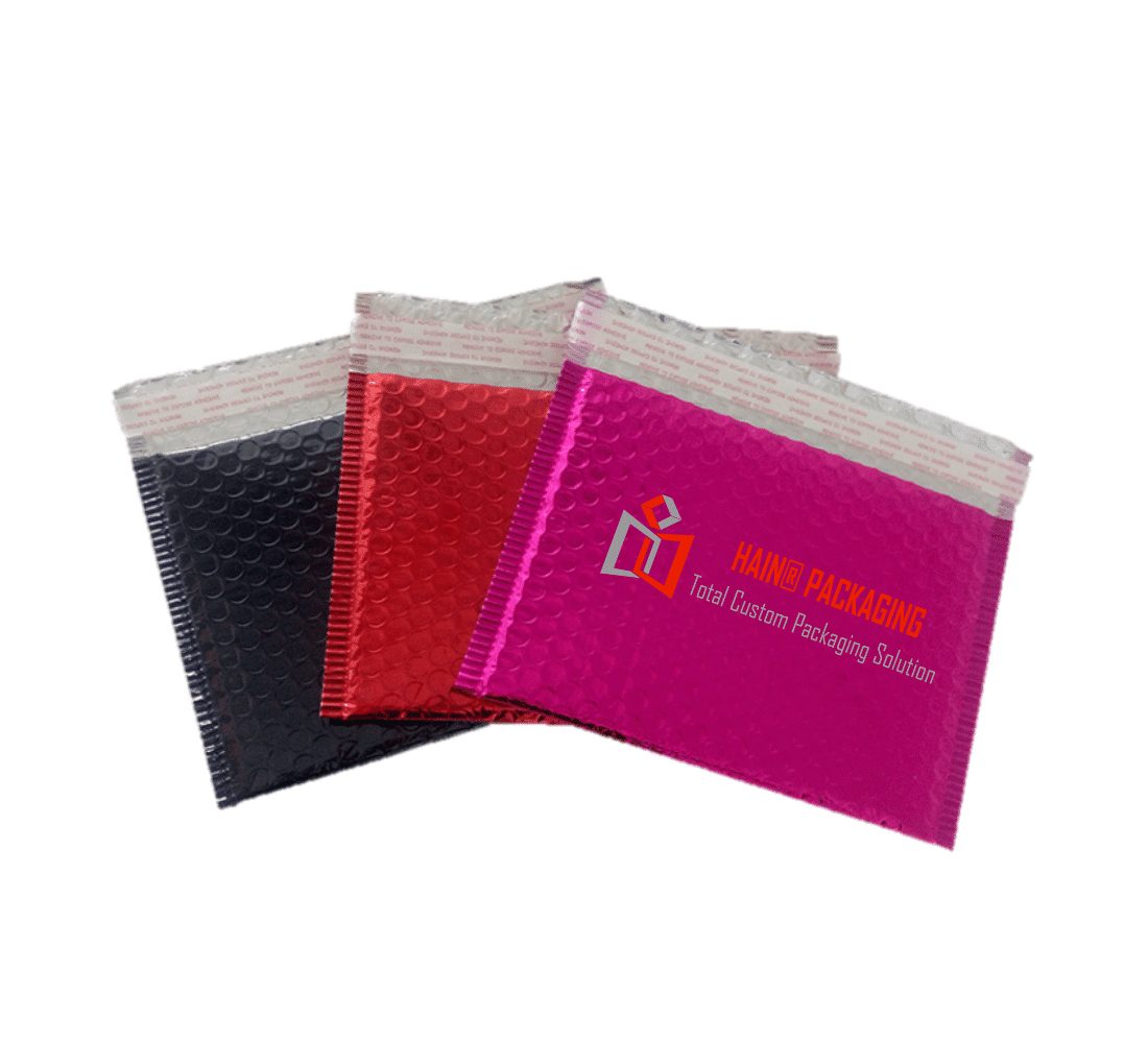 Three padded mailer envelopes in black, red, and bright pink, arranged diagonally. The pink envelope features a logo and text reading "HAIN® PACKAGING Total Custom Packaging Solution". All envelopes have a silver peel-and-stick closure strip.