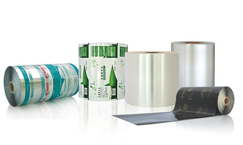 Five rolls of flexible packaging film are displayed on a white background. One roll lies horizontally on the left with blue and silver graphics. Three rolls stand vertically in the center and right, two appearing clear and one with green graphics. A fifth roll lies horizontally in the foreground with black and gold graphics.