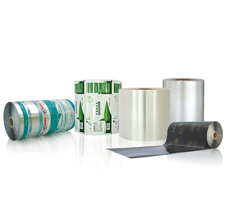 Five rolls of flexible packaging film are displayed on a white background. One roll lies horizontally on the left with blue and silver graphics. Three rolls stand vertically in the center and right, two appearing clear and one with green graphics. A fifth roll lies horizontally in the foreground with black and gold graphics.