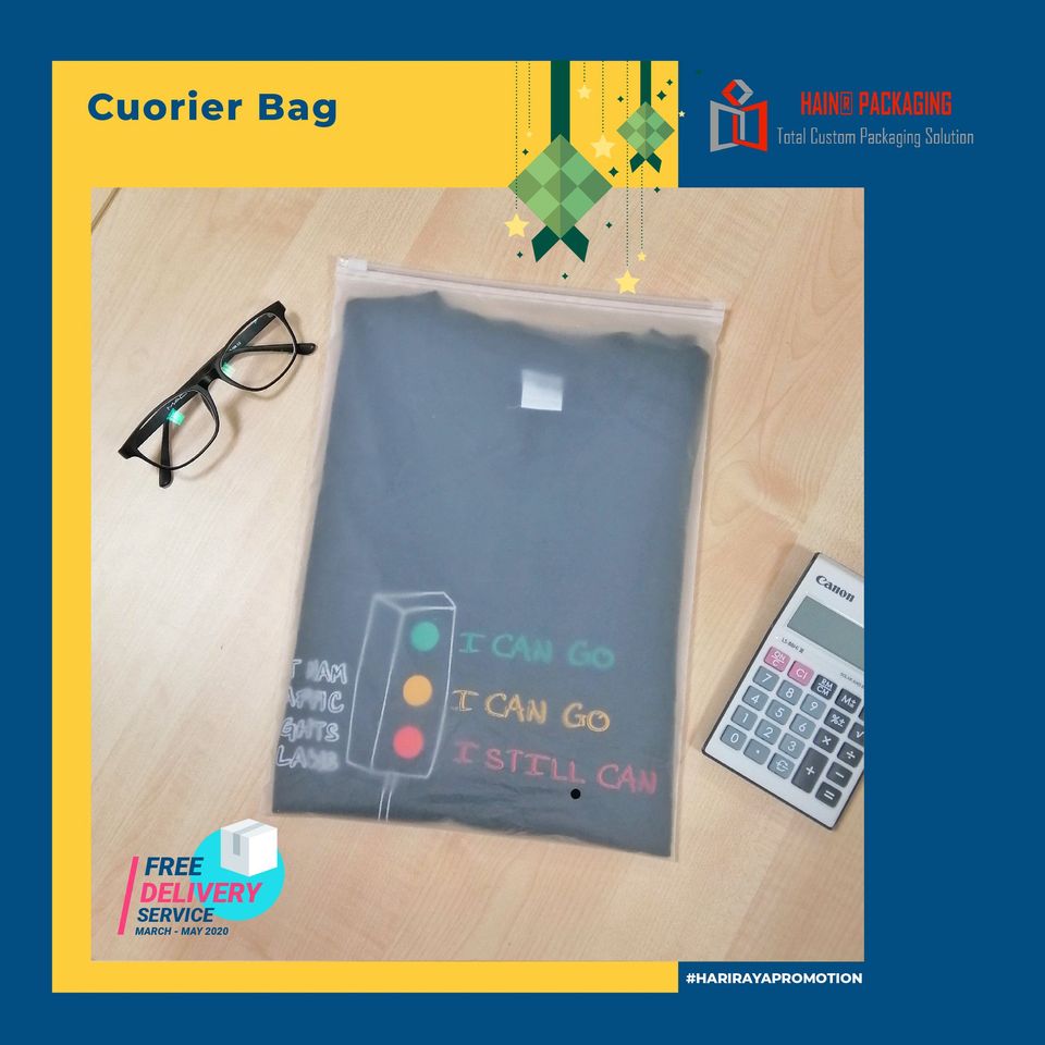 A clear courier bag containing a dark t-shirt with a traffic light graphic and text. A pair of glasses and a calculator are placed next to the bag on a wooden surface.
