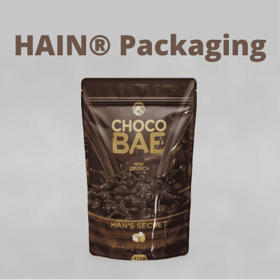 A dark brown stand-up pouch with a resealable closure, featuring gold lettering that reads 'CHOCO BAE' and 'HAN'S SECRET'. The pouch is filled with small, dark chocolate pieces.