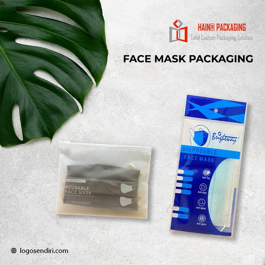 Two packages of face masks are displayed on a textured surface next to a large green leaf. One package is a translucent resealable pouch containing dark reusable masks. The other is a blue and white printed pouch with disposable masks visible inside.