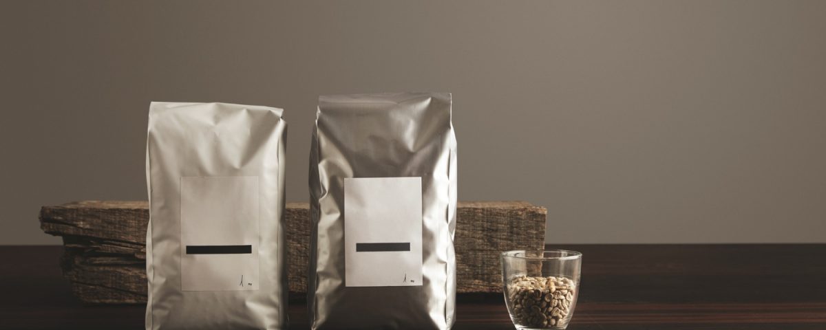 Two silver foil bags with white labels and black horizontal lines are displayed on a dark wooden surface. A small glass cup filled with coffee beans sits to the right of the bags.