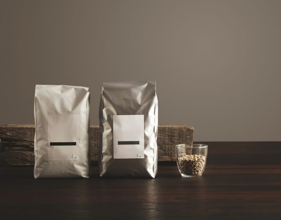 Two silver foil bags with white labels and black horizontal lines are displayed on a dark wooden surface. A small glass cup filled with coffee beans sits to the right of the bags.