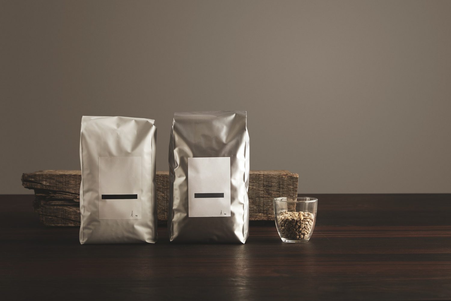 Two silver foil bags with white labels and black horizontal lines are displayed on a dark wooden surface. A small glass cup filled with coffee beans sits to the right of the bags.