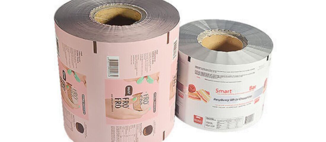 Two rolls of printed packaging film on white background. The larger roll on the left is pink with graphics and text. The smaller roll on the right is white with graphics and text.