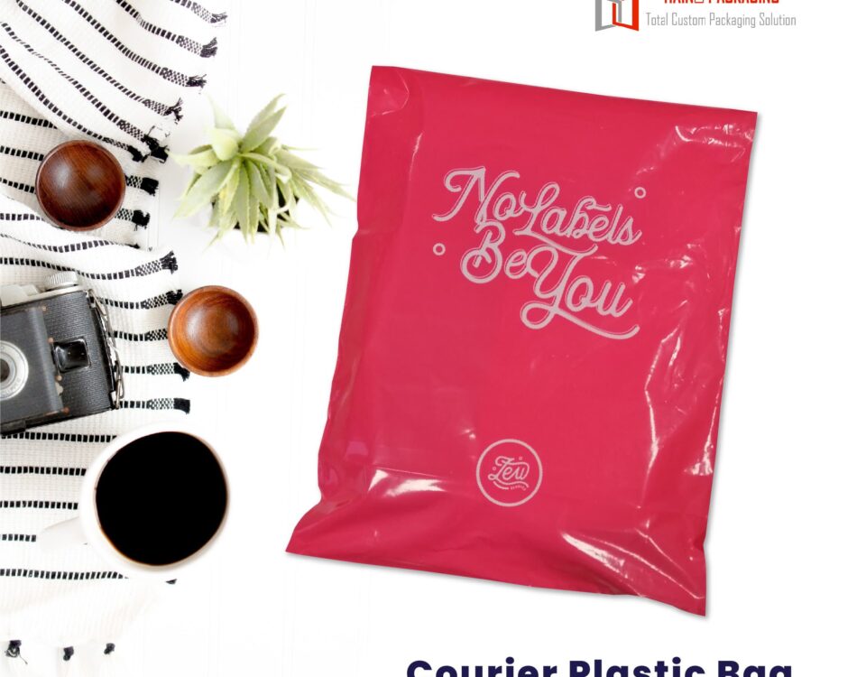 A bright pink, glossy courier mailer bag with white text that reads 'No Labels Be You' and a circular logo. The bag is shown flat against a white background, with a small plant, wooden bowls, a camera, and a cup of coffee to the left.