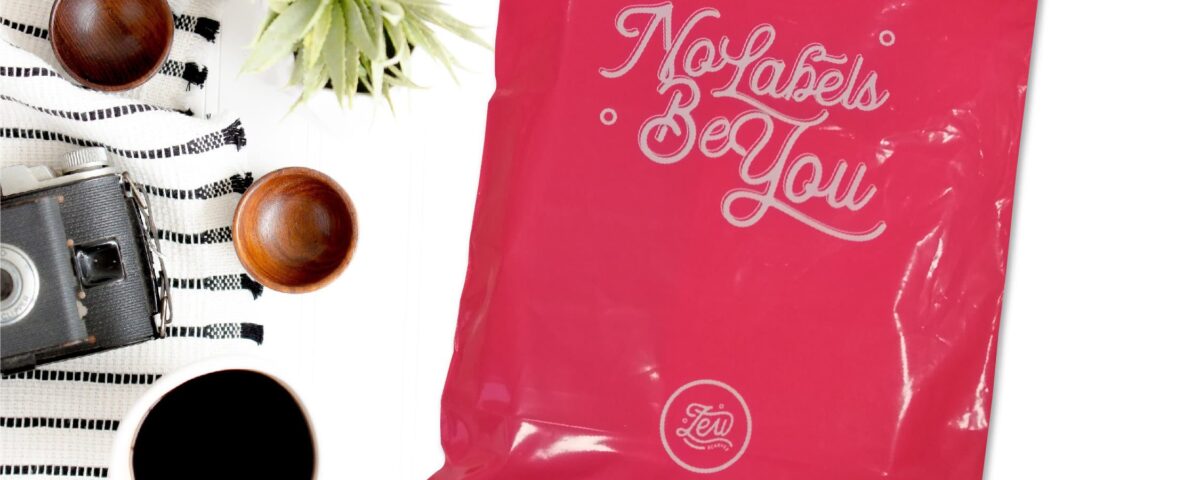 A bright pink courier mailer bag with white script text reading 'No Labels Be You' and a circular logo. The bag is shown flat on a white surface with a striped cloth, wooden bowls, a camera, and a cup of coffee.