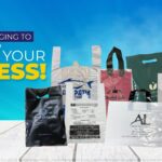 A collection of various plastic shopping bags with handles, displaying different colors, graphics, and printed text, arranged on a wooden surface against a blue sky background.