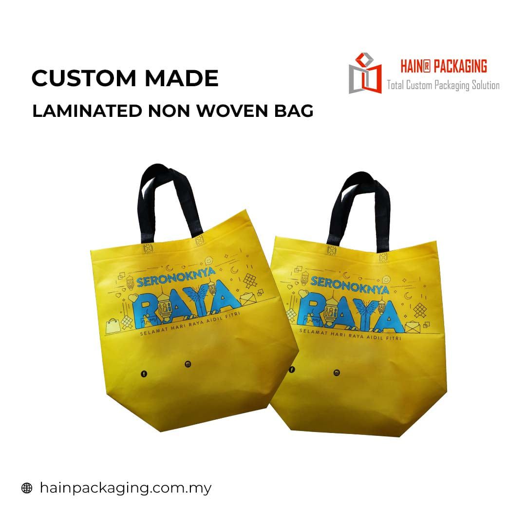 Two yellow laminated non-woven tote bags with black handles, each featuring a colorful graphic with the word "RAYA" and decorative elements, and text reading "SERONOKNYA" and "SELAMAT HARI RAYA AIDIL FITRI".