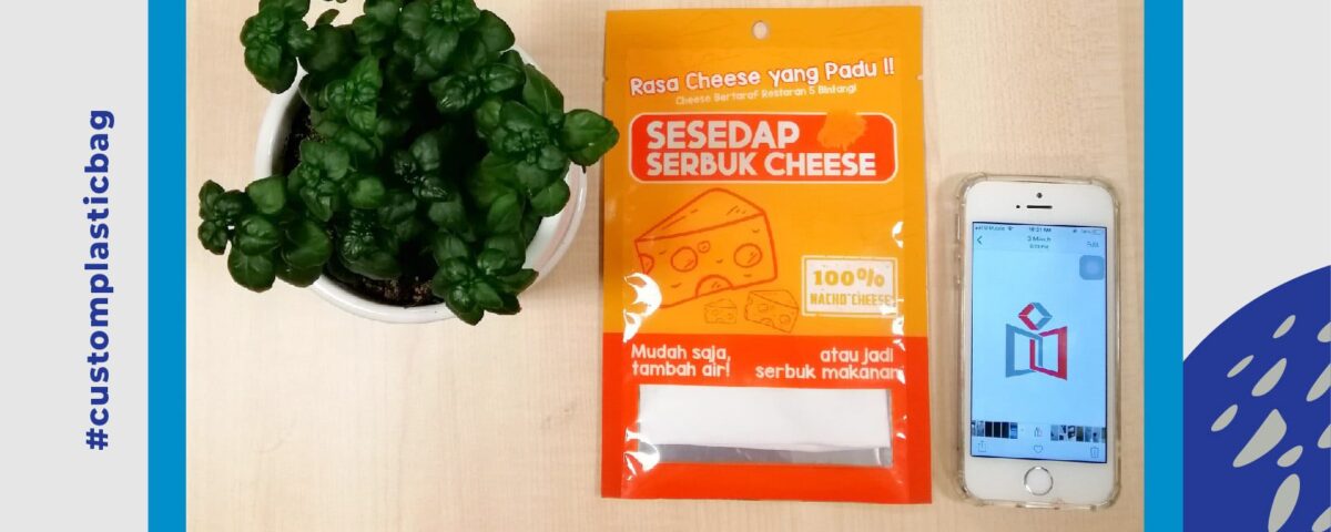 A bright orange and yellow three-side seal plastic bag for cheese powder seasoning, placed next to a potted plant and a smartphone displaying a logo.
