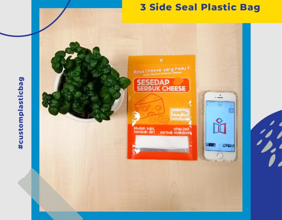 A bright orange and yellow three-side seal plastic bag for cheese powder seasoning, placed next to a potted plant and a smartphone displaying a logo.