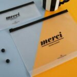 Two translucent document pouches with black zippers. One pouch is mostly clear with black text that reads 'merci' and 'SECOND MANSION'. The other pouch is half clear and half orange, also with black text reading 'merci' and 'SECOND MANSION'.