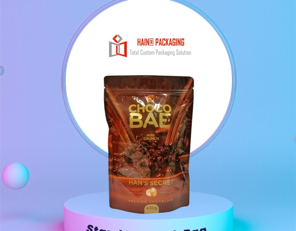 A brown stand-up pouch bag with a glossy finish, featuring a graphic of chocolate and text that reads 'CHOCO BAE MINI CRUNCH' and 'HAN'S SECRET PREMIUM CHOCOLATE 430g'. The bag is displayed on a light blue podium against a gradient background.