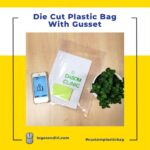 A clear plastic bag with gusseted sides lies open on a wooden surface. Inside are a white paper with green text and graphics, a small green notepad, and a smartphone displaying an app icon.