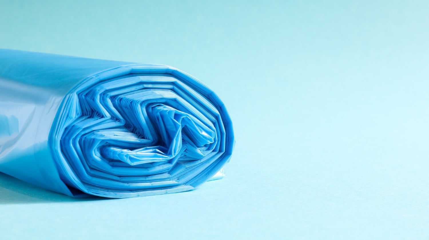 A close-up, side view of a roll of bright blue bags, tightly wound and resting on a light blue surface against a matching background.