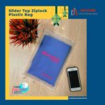 A clear slider top ziplock plastic bag containing a folded blue garment with pink text, placed on a wooden surface next to a smartphone and a decorative plant.