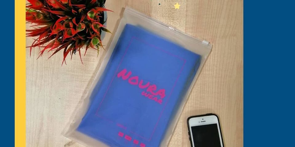 A clear slider top ziplock plastic bag containing a folded blue garment with pink text, placed on a wooden surface next to a smartphone and a decorative plant.
