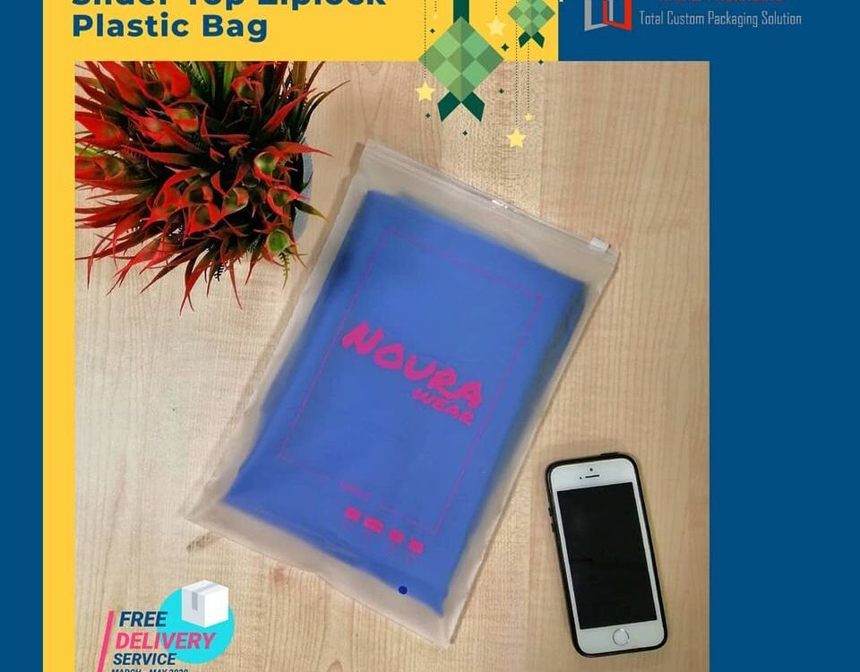 A clear slider top ziplock plastic bag containing a folded blue garment with pink text, placed on a wooden surface next to a smartphone and a decorative plant.