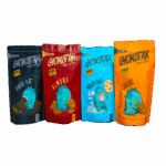 Four stand-up pouches of chocolate chip cookies, each with a different color and cartoon dinosaur graphic. From left to right: dark blue with dark oat flavor, red with almond flavor, light blue with milky suji flavor, and orange with classic flavor.