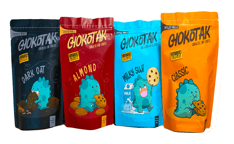 Four stand-up pouches of chocolate chip cookies, each with a different color and cartoon dinosaur graphic. From left to right: dark blue with dark oat flavor, red with almond flavor, light blue with milky suji flavor, and orange with classic flavor.