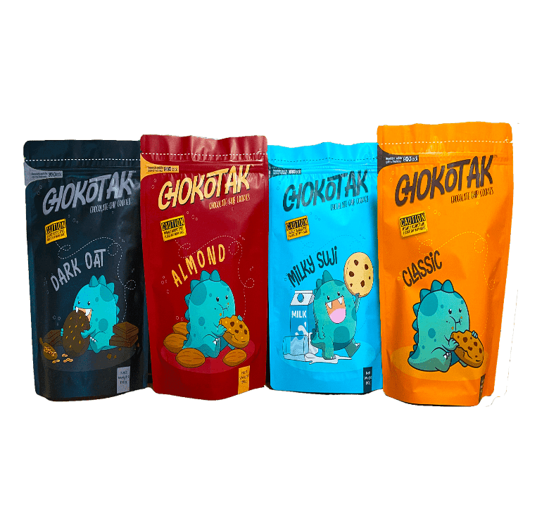 Four stand-up pouches of chocolate chip cookies, each with a different color and cartoon dinosaur graphic. From left to right: dark blue with dark oat flavor, red with almond flavor, light blue with milky suji flavor, and orange with classic flavor.